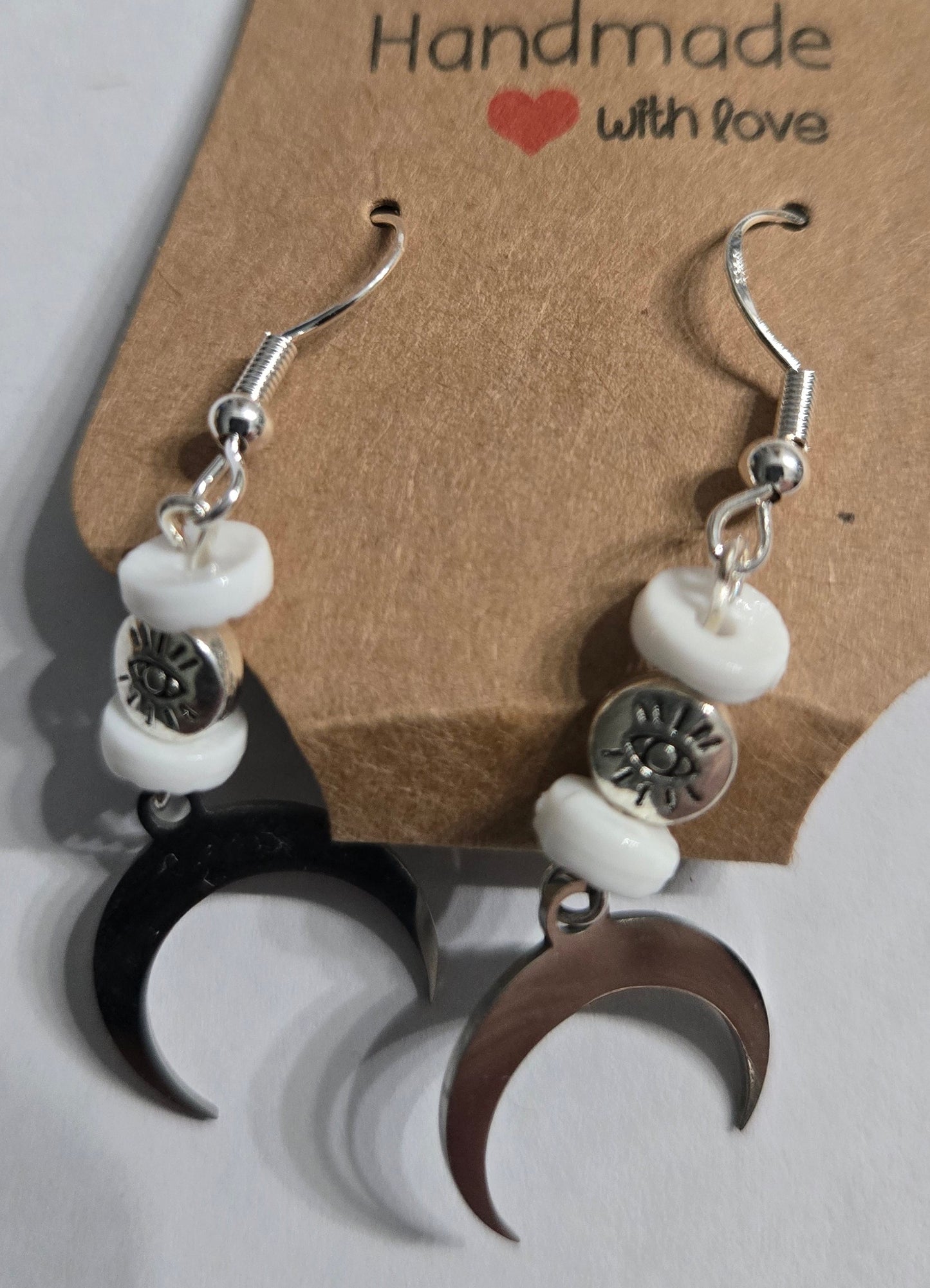 Handmade earrings with silver metallic crescent moon charms, accented with white glass spacer and silver in color "All Seeing Eye" beads, and crafted on sterling silver earring facets.